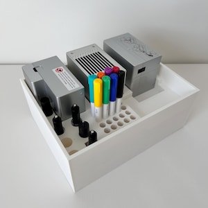 Puede incluir: Una caja organizadora de madera blanca que contiene dispositivos electrónicos plateados, objetos cilíndricos negros y marcadores de colores. Los marcadores incluyen tonos de azul, verde, amarillo, naranja, morado y negro. La caja está sobre una superficie blanca.