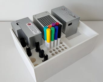 May include: A white wooden organizer box containing silver electronic devices, black cylindrical objects, and colorful markers. The markers include shades of blue, green, yellow, orange, purple, and black. The box is on a white surface.