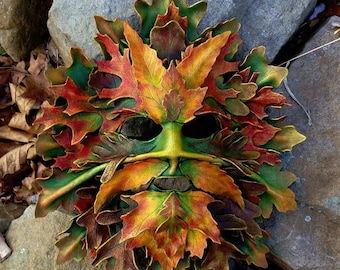 May include: A green, brown, and orange leather mask with a leafy design. The mask is shaped like a face with a long nose and large eyes. The leaves are arranged in a pattern that resembles a tree. The mask is made of leather and is decorated with gold paint.