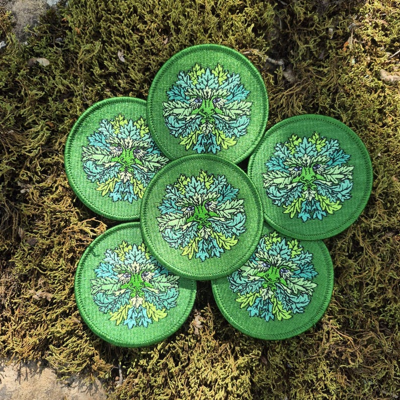 Peut inclure: Six &eacute;cussons verts circulaires avec un motif d'Homme Vert. La figure centrale pr&eacute;sente un visage avec des d&eacute;tails de feuilles dans des tons de vert et de bleu. Les &eacute;cussons sont dispos&eacute;s sur un lit de mousse verte.