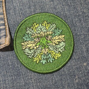 Peut inclure: Un &eacute;cusson rond et vert brod&eacute; repr&eacute;sentant l'Homme Vert. Le motif comprend un visage avec une barbe et une couronne de feuilles, dans des tons de vert et de jaune. L'&eacute;cusson est sur un fond en denim bleu.