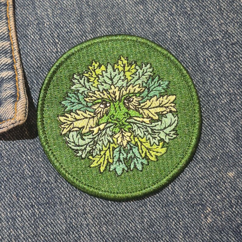 Peut inclure: Un &eacute;cusson rond et vert brod&eacute; repr&eacute;sentant l'Homme Vert. Le motif comprend un visage avec une barbe et une couronne de feuilles, dans des tons de vert et de jaune. L'&eacute;cusson est sur un fond en denim bleu.