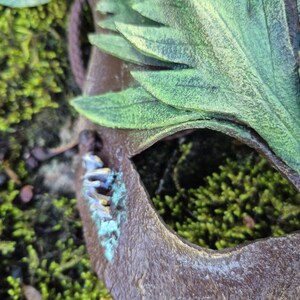 May include: A close-up of a brown mask with green leaf accents. The mask has a cut-out eye and is surrounded by green moss. The mask has a weathered appearance with hints of blue and gold.