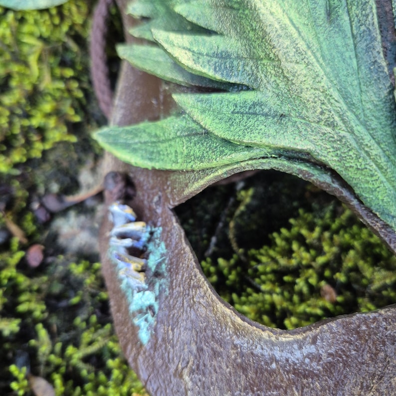 May include: A close-up of a brown mask with green leaf accents. The mask has a cut-out eye and is surrounded by green moss. The mask has a weathered appearance with hints of blue and gold.