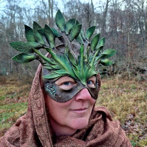 May include: A handcrafted, green and brown leather mask with a leafy branch design. The mask features detailed leaves and branches, creating a woodland or forest theme. The mask is worn by a person with a brown scarf.