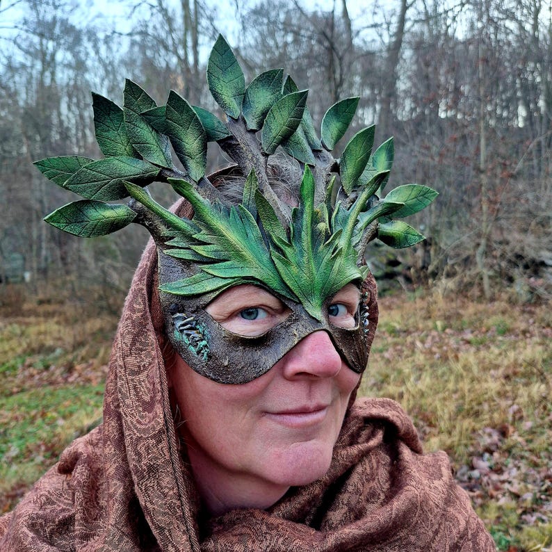 May include: A handcrafted, green and brown leather mask with a leafy branch design. The mask features detailed leaves and branches, creating a woodland or forest theme. The mask is worn by a person with a brown scarf.