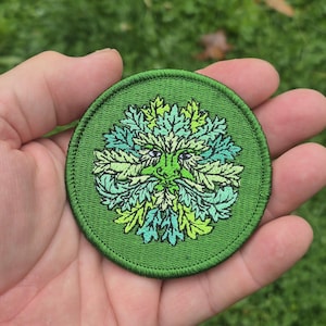 Peut inclure: Un &eacute;cusson rond et vert brod&eacute; repr&eacute;sentant l'Homme Vert. Le motif comprend un visage avec des traits de feuilles dans des tons de vert et de vert clair, avec de petits yeux violets. L'&eacute;cusson a une bordure verte.