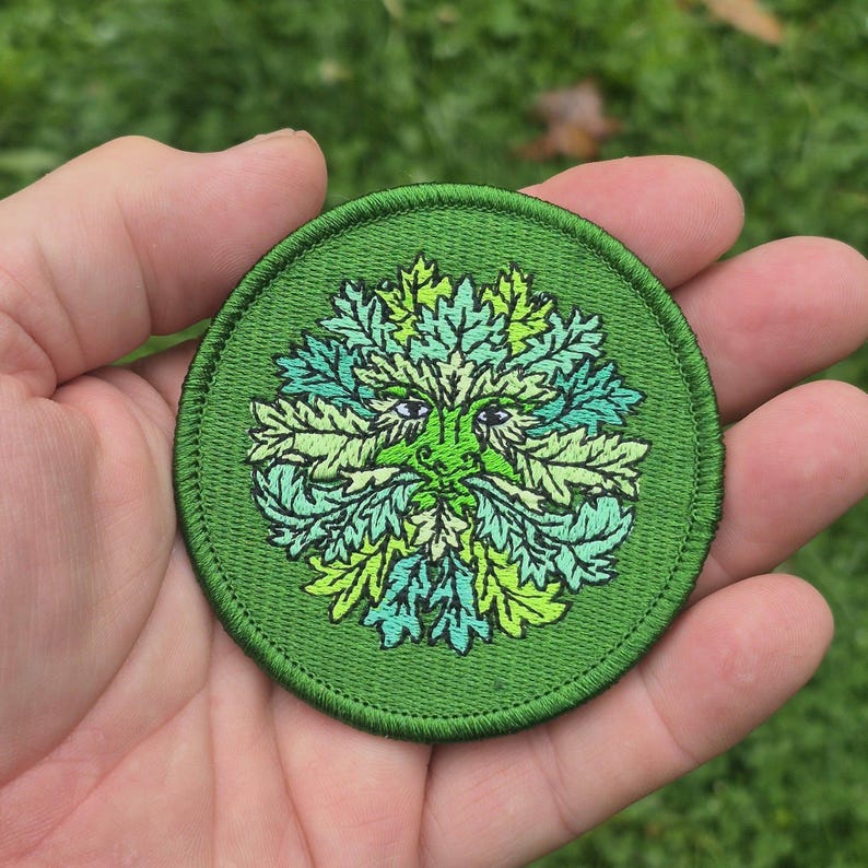 Peut inclure: Un &eacute;cusson rond et vert brod&eacute; repr&eacute;sentant l'Homme Vert. Le motif comprend un visage avec des traits de feuilles dans des tons de vert et de vert clair, avec de petits yeux violets. L'&eacute;cusson a une bordure verte.