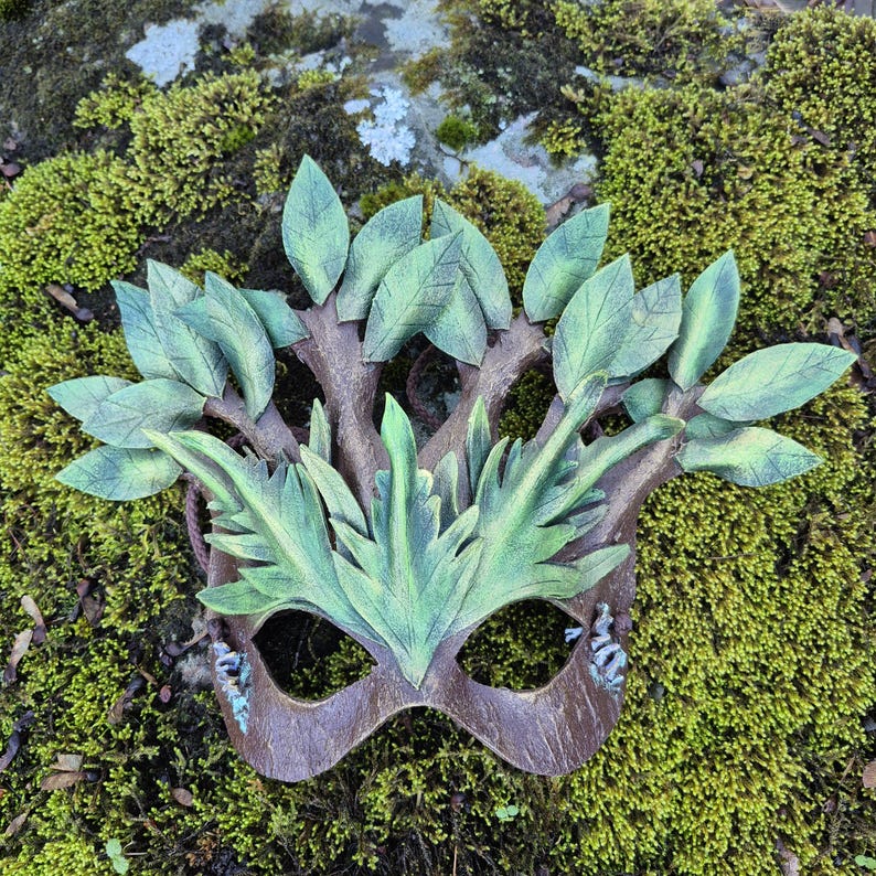May include: A handcrafted mask with a nature-inspired design. The mask features a brown base with green leaves and branches, creating a woodland aesthetic. The mask is designed with eye cutouts and is set against a mossy background.