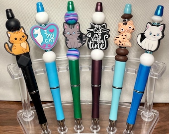 May include: A collection of six decorative pens with cat-themed toppers. Each pen features a different cat design, including orange, gray, and white cats, and the text "I My Cat" and "My Cat My Valentine". The pens are displayed in a clear acrylic holder.