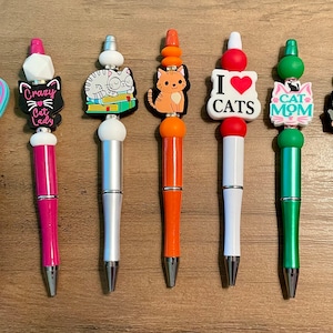 May include: Set of ten colorful pens with cat-themed toppers. The toppers include a cat reading a book, a cat with a pizza, a cat with a heart, a cat with a crown, and a cat with a crown.