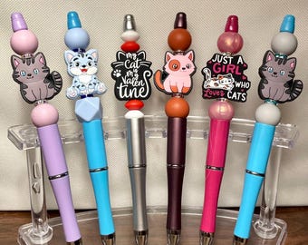 May include: A collection of decorative pens with cat-themed toppers. Each pen features a different color barrel and a unique cat design or phrase, including "My Cat is My Valentine" and "Just a Girl Who Loves Cats."