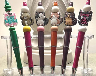 May include: A set of six decorative pens with cat-themed toppers. The pens have various colors, including green, pink, purple, and orange. Some pens feature cat-shaped charms, while others have the phrases "Cat Mom" and "Crazy Cat Lady."