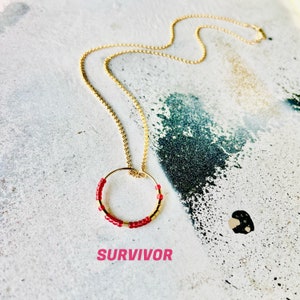 Puede incluir: Un collar de cadena dorada con un colgante circular de cuentas rojas. El colgante es un círculo simple con cuentas rojas alrededor del borde. El collar está sobre un fondo blanco con la palabra "SURVIVOR" escrita en rosa debajo del colgante.