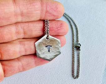 Puede incluir: Un collar con colgante hexagonal de tono plateado que presenta una superficie martillada y texturizada con un pequeño icono de tornillo oscuro estampado en el centro. El colgante cuelga de una cadena de metal plateado fina y delicada con un pequeño conector circular.