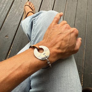 Puede incluir: Una pulsera plateada con un colgante circular y una correa de cuero marrón. El colgante tiene una superficie texturizada y un pequeño recorte. La pulsera se abrocha con un cierre plateado.