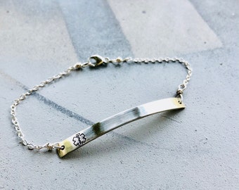 以下が含まれることがあります： つや消し金属仕上げのシルバーカラーの医療IDブレスレット。ブレスレットは、刻印された医療シンボルと繊細なチェーンが付いた長方形のプレートが特徴です。留め金が見え、灰色の背景に設定されています。