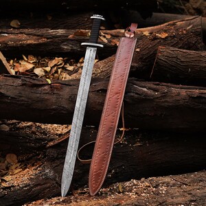 May include: A silver sword with a textured blade and a black handle rests beside a brown leather sheath. The sword has a crossguard and a rounded pommel. The sheath is detailed with stitching and a leather loop.