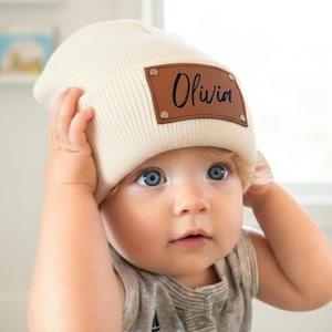 Puede incluir: Gorro color crema con un parche de cuero marrón que dice "Olivia" en escritura negra. El gorro es de canalé y tiene un borde doblado. El bebé de la imagen lleva un body gris.