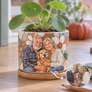 May include: A cylindrical ceramic planter featuring a colorful illustration of an older couple and a golden retriever dog, surrounded by a large floral pattern. The planter sits on a circular wooden base and contains a green Pilea plant.