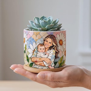 May include: A cylindrical ceramic planter featuring a colorful mosaic-style illustration of a person holding a baby, surrounded by white daisy flowers. A green succulent plant sits inside, and the pot rests on a circular wooden base.