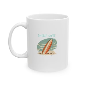 May include: White ceramic mug featuring a surfboard design with the text "SURF LIFE". The illustration shows a surfboard in the ocean with waves and sand. The mug has a curved handle.