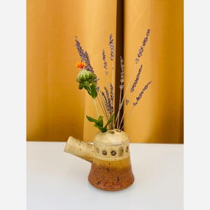 May include: A ceramic vase with a spout, featuring a gradient of colors from beige to brown. The vase holds dried lavender and a single orange flower. The background is a solid mustard yellow.
