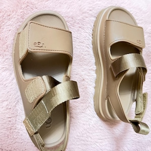 May include: A pair of beige sandals with adjustable straps. The sandals have a thick sole and the brand name "UGG" is visible on the straps. The sandals are designed for comfort and casual wear.