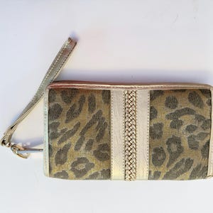 May include: A rectangular wallet with a gold wrist strap. The wallet features a leopard print design in shades of brown and gold, with a central vertical stripe of white and braided gold material. The edges are trimmed in gold.