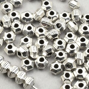 May include: Silver-coloured, faceted beads with a hexagonal shape. The beads are scattered across a light grey surface, some strung on a thin wire. The beads have a metallic sheen, suitable for jewellery making.