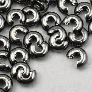 May include: Close-up of a pile of silver-colored crimp beads. These small, C-shaped beads are used in jewelry making to secure clasps and other components. The beads are scattered on a light-colored surface.