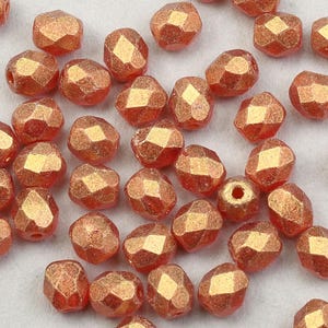 May include: A collection of small, faceted glass beads in a copper color. The beads have a metallic sheen and are scattered on a white surface.