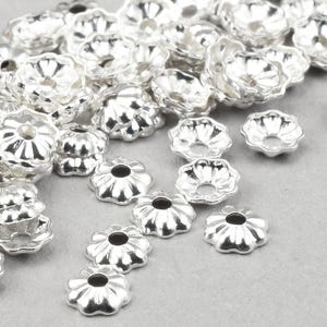 May include: A collection of small, silver-colored bead caps with a flower-like design. The caps are approximately 3.5mm in size and are scattered across a light gray surface. The text at the bottom of the image reads: "These 3.5mm caps are very small!"