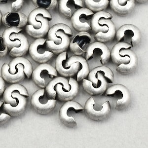 May include: Close-up of a collection of silver-colored crimp beads. The beads are small, curved, and have a split opening. They are scattered on a light gray surface, ideal for jewelry making.
