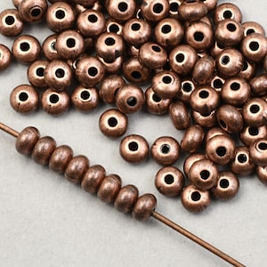 May include: A pile of small, round, copper-colored beads with a matte finish. The beads are scattered on a gray surface. A thin wire is threaded with a string of the same beads.