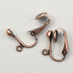 May include: Two antique copper-colored clip-on earring findings. Each finding has a curved, hinged clip with a textured pad and a small loop. The findings are designed for attaching to earrings.