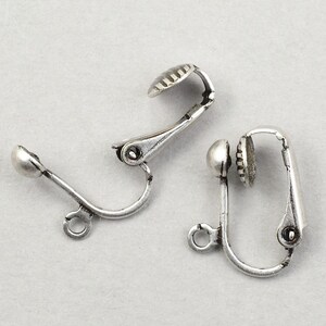 May include: Two silver-colored clip-on earring findings. Each finding has a curved arm with a small loop, a rounded end, and a textured, oval-shaped clip. The findings are designed for jewelry making.