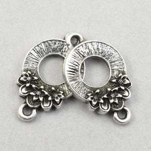 May include: Two silver-toned metal jewelry components. Each component features a circular design with a textured surface and a cluster of small flowers. A loop is at the bottom for attachment. Ideal for jewelry making.