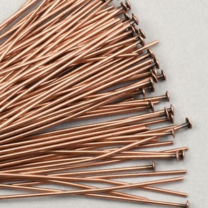 May include: A pile of copper-colored head pins, each with a flat, round head and a long, slender shaft. The pins are arranged in a dense, overlapping pattern, with some extending towards the viewer. The background is a light gray.
