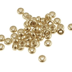May include: A pile of gold-colored metal beads, each with a round hole in the center. The beads are scattered on a white background.