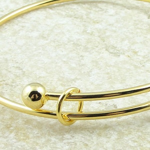 May include: A gold-colored bangle bracelet with two small, round, gold-colored beads on either side of a decorative clasp. The bracelet is made of a thin, curved metal band.
