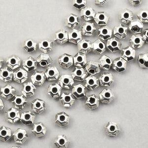 May include: A scattering of small, silver-coloured, geometric beads. Each bead has multiple facets and a central hole. They are spread across a light grey surface, highlighting their intricate design and metallic sheen. Suitable for jewellery making.