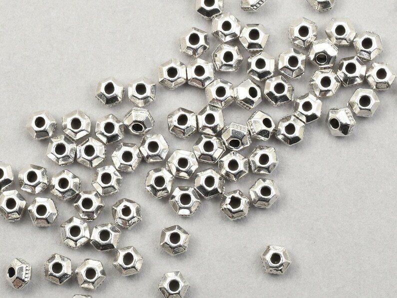 May include: A scattering of small, silver-coloured, geometric beads. Each bead has multiple facets and a central hole. They are spread across a light grey surface, highlighting their intricate design and metallic sheen. Suitable for jewellery making.
