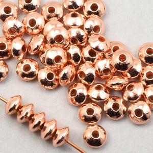 May include: A pile of rose gold metal beads with a hole in the center. The beads are round and have a slightly domed shape. A strand of the beads is strung on a wire.