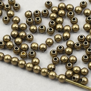 May include: A collection of small, round, brass-colored beads with a hole through the center. Some beads are strung on a thin, gold-colored wire. The beads are scattered across a white surface, suitable for jewelry making or crafts.