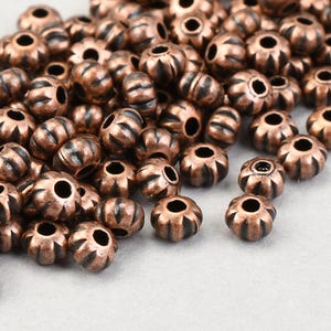 May include: A collection of small, copper-colored metal beads. Each bead has a ribbed, barrel-like shape with dark accents and a central hole. The beads are scattered, showcasing their detailed design.