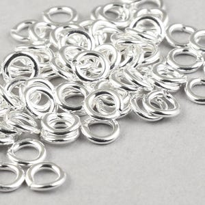 May include: A pile of small, shiny, silver-colored jump rings. These round metal rings are open and ready to be used in jewelry making or other craft projects. The rings are scattered on a light gray surface.