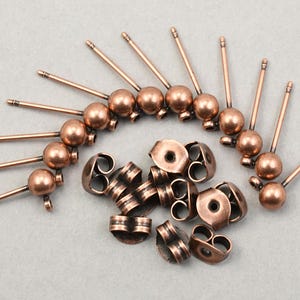 May include: Copper-colored earring posts with ball-shaped accents and matching earring backs. The posts have a slender design, and the earring backs are butterfly style. The components are arranged on a light gray surface.