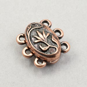May include: An oval-shaped copper-colored clasp with a floral design in the center. The clasp has six loops around the oval. The floral design is a stylized leaf and stem pattern.