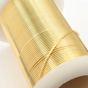 May include: A spool of gold-colored wire, tightly wound around a white plastic core. The wire is smooth and reflective, catching the light. This item is likely used for jewelry making or crafting.